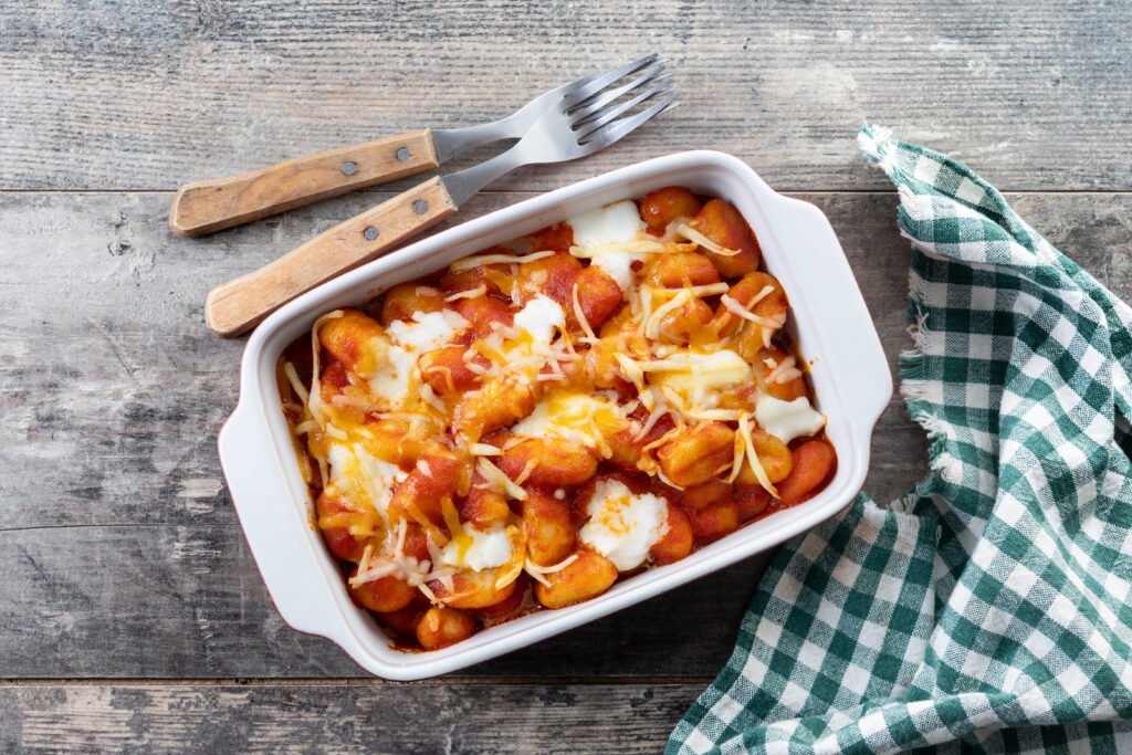 Baked Gnocchi alla Sorrentina in a white casserole dish on a rustic wooden table with a checkered napkin and wooden cutlery.